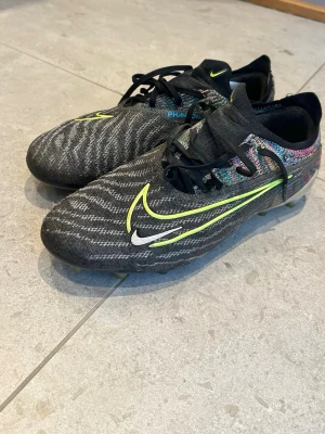 Nike Phantom GX Elite fotbollsskor AG  - Säljer ett par Nike Phantom GX Elite fotbollsskor i svart med neonfärgade detaljer och vit logga. Strl 38,5. Skorna har snörning och en modern, strömlinjeformad design. Ovandelen är tillverkad i syntetmaterial som kallas för gripknit för bra bollkontroll. Skorna har en AG platta som rekommenderas att spelas på konstgräs planer men funkar bra på gräs också. Medföljer originalpåse. Kommer från djur- och rökfritt hem.