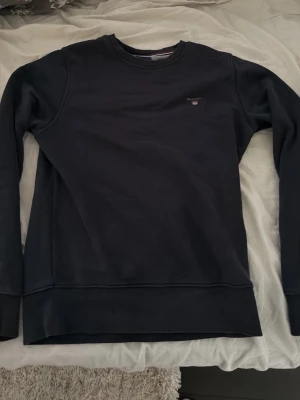 Mörkblå sweatshirt från Gant - En stilren mörkblå sweatshirt från Gant med rund halsringning och diskret logga broderad på bröstet. Perfekt för en clean och avslappnad look. Perfekt skick i storlek S