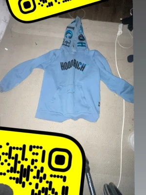 Blå HOODRICH hoodie med tryck - Säljer en ljusblå hoodie från HOODRICH med svart och blått tryck på bröstet och textdetaljer på huvan. Hoodien har en klassisk känguruficka framtill och en stor huva med tryck. Perfekt för dig som gillar streetwear och vill sticka ut.