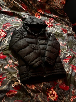 Svart dunjacka Peak Performance - Snygg svart dunjacka från Peak Performance med huva och dragkedja framtill. Jackan har diskret logga broderad på bröstet och är perfekt för kalla vinterdagar. Puffig design och klassisk look som funkar till det mesta.