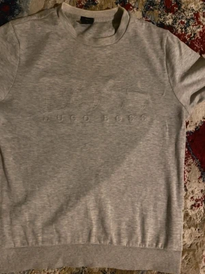 Hugo Boss tröja  - Snygg ljusgrå kortärmad tröja från Hugo Boss med diskret broderad logga framtill. Tröjan har rund halsringning och ribbade muddar vid ärmslut och nederkant. Perfekt för en clean och stilren look.