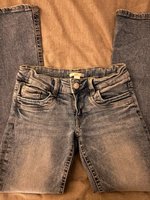 Blå slitna bootcut jeans - Säljer ett par blå bootcut jeans med snyggt slitna detaljer och klassiska fem fickor. Materialet är denim med en mjuk känsla och tvättad look som ger en avslappnad vibe. Jeansen har justerbar midja