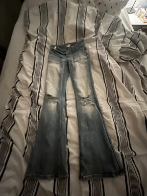 Blå bootcut jeans från Gina tricot  - Säljer ett par ljusblå bootcut jeans med slitningar över knäna och broderade detaljer på bakfickorna. Jeansen har en klassisk femficksmodell och är tillverkade i denim med tvättad look. Perfekta för en avslappnad och trendig stil. Från Gina tricot.