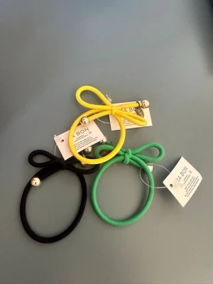 Färgglada hårsnoddar med rosett - Säljer ett set med hårsnoddar från JABON i svart, gul och grön. Varje snodd har en söt rosett och små silvriga bollar som dekoration. Perfekta för att piffa upp din frisyr och hålla håret på plats. Tillverkade i mjukt elastiskt material.