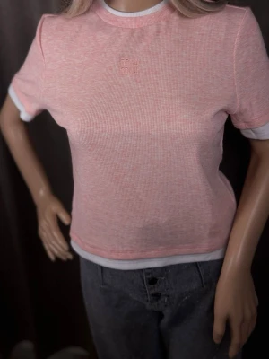 Rosa ribbad t-shirt från Miu Miu - Säljer en ljusrosa ribbad t-shirt från Miu Miu med broderad logga framtill. T-shirten har korta ärmar och vita kontrastdetaljer vid halsringning och ärmslut. Perfekt för dig som gillar en clean och trendig look.