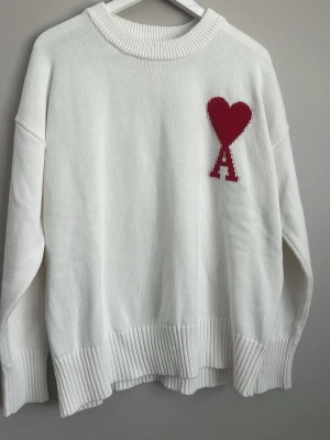 Vit stickad tröja Ami Paris hjärta - Vit oversized stickad tröja från Ami Paris med röd hjärtdetalj och bokstaven 'A' på bröstet. Tröjan har bred ribbstickad kant vid hals, ärmslut och nederkant. Perfekt för en trendig och avslappnad look.