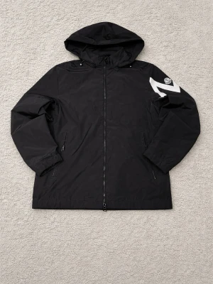 Svart Moncler vindjacka med huva - Svart vindjacka från Moncler med huva och dragkedja framtill. Jackan har två sidofickor med dragkedja och en vit grafisk detalj på ena ärmen samt Moncler-logga. Tillverkad i ett lätt, vattenavvisande material som passar perfekt för blåsiga dagar och sommarkvällar( pris kan diskuteras)