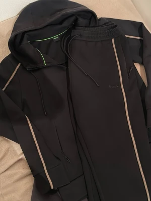 Ny Svart dress från BOSS med beige detaljer - Snygg svart dress från BOSS med utsvängda byxor. dragkedja och beige ränder längs ärmarna. Huva med snörning och diskret BOSS-logga på bröstet. Insidan av huvan har en grön detalj. Tillverkad i mjukt och stretchigt material för extra komfort.