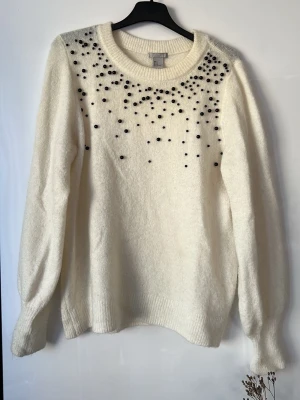 Vit stickad tröja med pärlor H&M - Säljer en krämvit stickad tröja från H&M med svarta pärlor över axlar och bröst. Tröjan har rund halsringning, långa ärmar och ribbade muddar. Finns en beige fläck vid ärmen men inget som syns. Pris kan diskuteras