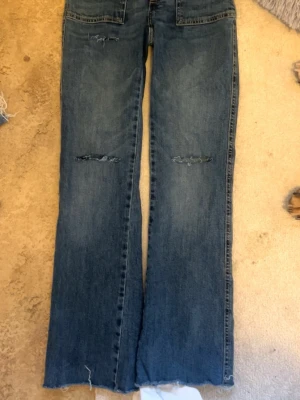 Blå bootcut jeans med slitningar - Säljer ett par blå bootcut jeans med slitna detaljer vid knäna och råa, fransiga benslut. Jeansen har klassisk femficksdesign och är gjorda i denim. Perfekta för dig som gillar en avslappnad och trendig stil.