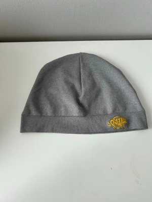 Syna skull cap - Snygg grå mössa i mjukt tyg med gul broderad textdetalj framtill. Enkel och stilren design som passar till de flesta outfits. Perfekt accessoar för kyliga dagar.