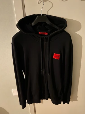 Svart hoodie från Hugo med dragkedja - Säljer en svart hoodie från Hugo med dragkedja framtill och justerbar huva. På bröstet sitter en röd patch med Hugo-loggan. Hoodien har fickor på sidorna och är gjord i mjukt material som känns skönt mot huden. Perfekt för en avslappnad och stilren look.
