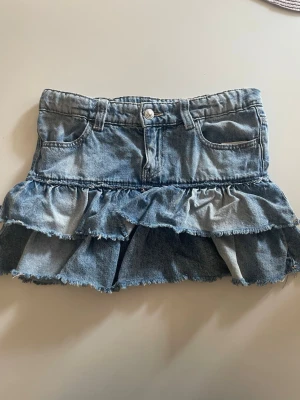 Blå jeanskjol med volanger - Supersöt blå jeanskjol med dubbla volanger och råa kanter i storlek 152. Kjolen har klassiska fickor fram samt hällor för skärp. Perfekt för dig som gillar en avslappnad men trendig stil. Tillverkad i denim med snygg tvättad look💙