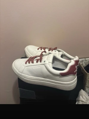 Skor  - Stilrena vita sneakers från Björn Borg  med vinröda detaljer på hälen och matchande vinröda skosnören. Klassisk låg modell med rund tå och vit sula. Perfekta för dig som vill ha en clean look med en twist.