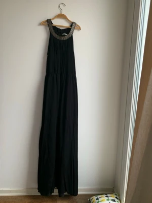 Svart långklänning från Copenhagen Luxe - Elegant svart långklänning från Copenhagen Luxe med glittriga pärldetaljer runt halsringningen. Klänningen har ärmlös design och ett mjukt, fallande tyg som ger en snygg siluett. Perfekt för speciella tillfällen när du vill sticka ut.