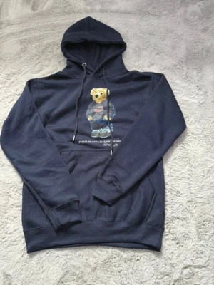Mörkblå Polo Bear hoodie Ralph Lauren - Mörkblå hoodie från Ralph Lauren med den ikoniska Polo Bear-trycket på bröstet. Hoodien har huva med dragsko och en stor magficka. Tillverkad i mjukt bomullsmaterial och har ribbade muddar vid ärmslut och nederkant. Perfekt för en avslappnad och stilren look.