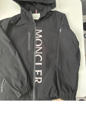 Svart vindjacka från Moncler - Snygg svart vindjacka från Moncler med stor vit logga längs dragkedjan framtill. Jackan har huva, bröstficka med dragkedja och elastiska muddar vid ärmslut och midja. Tillverkad i ett lätt, vattenavvisande material och har Moncler-märke på ärmen.