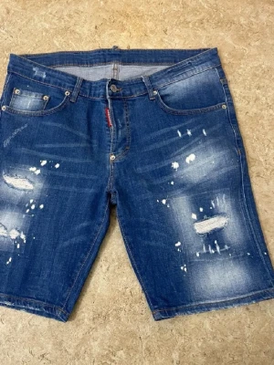 Blå distressed jeansshorts Dsquared2 - Säljer ett par blå jeansshorts från Dsquared2 med slitna detaljer och distressed look. Klassisk femficksmodell med knappgylf och bälteshällor. Shortsen har en rå och trendig känsla, perfekta för dig som gillar streetwear och denim.