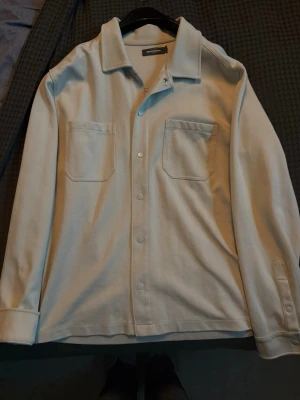 Vit overshirt från Dressmann - Vit overshirt från Dressmann med klassisk krage, två bröstfickor och knäppning framtill. Materialet är mjukt och har en slät finish. Perfekt att bära som lager över t-shirt eller skjorta. Enkel och stilren design som passar till många outfits.