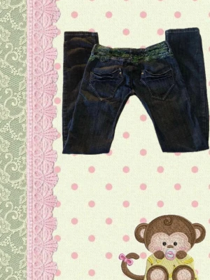Mörkblå jeans med 2000s/y2k stil - Snygga mörkblå jeans med unika gröna spetsdetaljer i midjan och dekorativa sömmar. Jeansen har klassiska fem fickor och knappgylf. Perfekta för dig som vill ha något extra i garderoben.