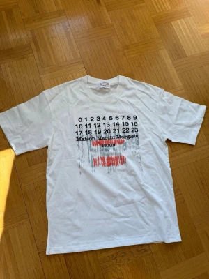 Maison Margiela vit t-shirt med tryck - Vit t-shirt från Maison Martin Margiela med ikoniskt siffertryck och röd text på bröstet. Klassisk passform med korta ärmar och rund hals. Tillverkad i mjuk bomull, perfekt för en clean och modern stil. Använd fåtal gånger, storlek L. Hör av er vid frågor☺️