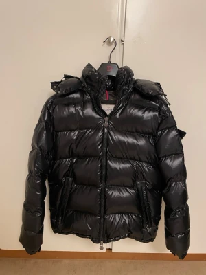 Moncler jacka  - Säljer en svart glansig dunjacka med huva och dragkedja framtill. Jackan har två stora fickor med dragkedja och en hög krage.