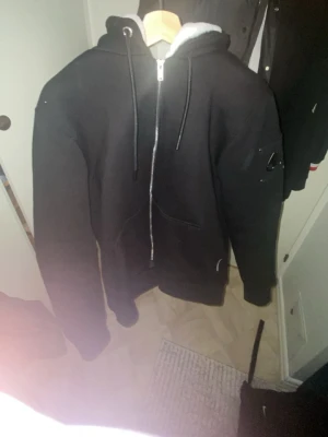 Svart hoodie från Moose Knuckles - Säljer en svart hoodie från Moose Knuckles med dragkedja, huva med snörning och mjukt foder. På ärmen sitter det ikoniska metall-logotypen på en patch med nitar. Hoodien har känguruficka och ribbade muddar. Perfekt för dig som gillar streetwear och snygga detaljer.