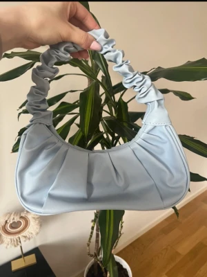 Ljusblå axelväska med rynkat handtag - Trendig ljusblå handväska i mjukt fejkläder med rynkade detaljer och ett scrunchie-inspirerat handtag. Väskan har en rundad form och veckad framsida som ger en cool och modern look. Perfekt accessoar för att lyfta din outfit.