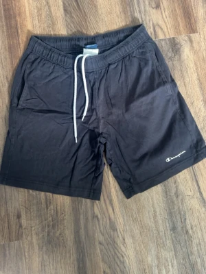 Svarta shorts från Champion - Svarta shorts från Champion med vit snörning i midjan och diskret logga på vänster ben. Tillverkade i mjuk bomull för skön känsla. Perfekta för chill dagar eller träning.