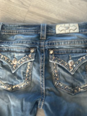 Blå slitna jeans med broderade fickor - Säljer ett par blå jeans från Miss Me med slitningar och hål på benen för en edgy look. Bakfickorna har snygga broderier och dekorativa knappar. Jeansen har bootcut-modell och detaljerade sömmar. Perfekta för dig som gillar statement-plagg!