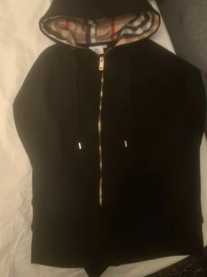 Burberry Hoodie - Snygg svart hoodie från Burberry med klassiskt rutigt foder i huvan och guldfärgad dragkedja. Hoodien har justerbara snören i huvan och diskret broderad logga på bröstet. Perfekt för dig som vill ha en stilren och exklusiv look.