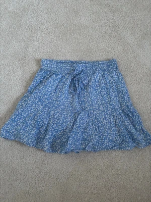 Blå blommig kjol - En jättefin blå blommig kjol med vita blommor från Pigalle i storlek M men passar även S. Kjolen har även insydda shorts och är i väldigt bra skick.