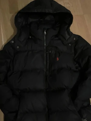 Svart dunjacka Polo Ralph Lauren - Säljer en svart dunjacka från Polo Ralph Lauren med huva och dragkedja framtill. Jackan har en bröstficka med dragkedja och den klassiska röda loggan på bröstet. Puffig modell som håller dig varm under kalla dagar.