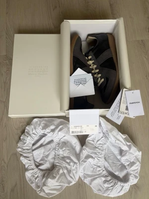 Maison Margiela Replica sneakers grå - Snygga Maison Margiela Replica sneakers i mörkgrå mocka och läder med beige skosnören och klassisk gummisula. Ikonisk design med diskret logga på plösen och stilrena detaljer. Perfekta för dig som gillar minimalistisk och trendig stil.