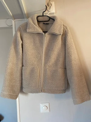 Beige teddyjacka med dragkedja - Mysig beige teddyjacka med hög krage och hel dragkedja framtill. Jackan har två stora fickor på framsidan och är tillverkad i fluffigt, mjukt teddy-material som håller dig varm. Perfekt för kyliga dagar och enkel att matcha med olika outfits.