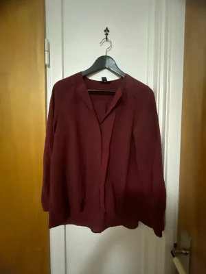 Vinröd blus med krage - Stilren vinröd blus med klassisk krage och dold knappslå. Blusen har lång ärm och ett mjukt, lätt tyg som faller snyggt. Perfekt att matcha med jeans eller kjol för en trendig look.