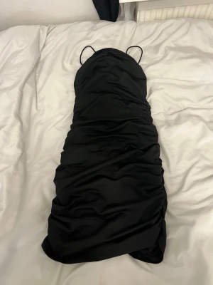 Svart omlottklänning med smala band - Snygg svart kortklänning med smala axelband och omlottdesign. Klänningen har rynkade detaljer längs sidorna och en djup v-ringning framtill. Perfekt för dig som vill ha en stilren och trendig look. Är väldigt stretchig så passar även som storlek S