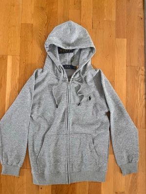Grå zip hoodie från Polo Ralph Lauren - Snygg grå hoodie med dragkedja från Polo Ralph Lauren. Klassisk huva med dragsko, två fickor framtill och den ikoniska svarta loggan broderad på bröstet. Tillverkad i mjuk bomullsblandning som känns skön mot huden. Perfekt för en chill och stilren look.