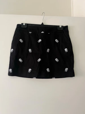 Svart kortkjol med dödskallemönster - Svart kortkjol med vita dödskallar broderade över hela tyget. Kjolen har en rak passform och sidofickor, perfekt för dig som gillar en edgy och unik stil. Materialet känns som bomull och kjolen är enkel att matcha med olika toppar.
