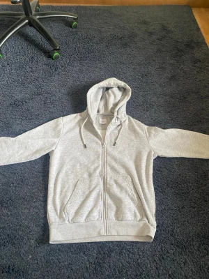Ljusgrå zip hoodie från Jack & Jones - Ljusgrå hoodie med dragkedja från Jack & Jones. Klassisk modell med huva och dragsnören, två stora fickor framtill och ribbade muddar vid ärmslut och nederkant. Tillverkad i mjukt sweatshirtmaterial för maximal komfort. Hör av er vid frågor eller funderingar 