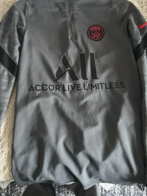 Grå PSG träningströja Nike - Snygg grå träningströja från Nike med PSG-logga på bröstet och svart 'Accor Live Limitless'-tryck på framsidan. Halvlång dragkedja framtill och röd Nike-logga på ärmen. Perfekt för fotbollsträning eller chill.