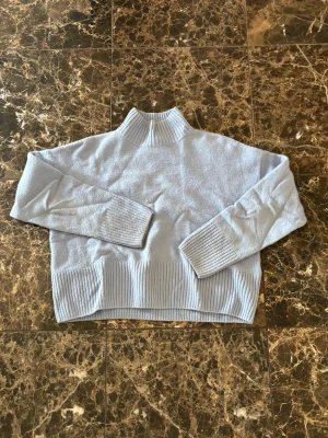 Ljusblå stickad polotröja - Mysig ljusblå stickad tröja med hög krage och breda ribbade muddar vid ärmslut och nederkant. Tröjan har en avslappnad passform och är perfekt för kyliga dagar. Materialet känns mjukt och bekvämt mot huden.