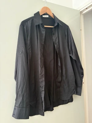 Svart skjorta från Why Denim - Svart skjorta från Why Denim med klassisk krage och knappar framtill. Skjortan har lång ärm med knappdetalj på ärmen och är tillverkad i ett slätt material som ger en stilren look. Perfekt för dig som gillar enkel och tidlös stil.