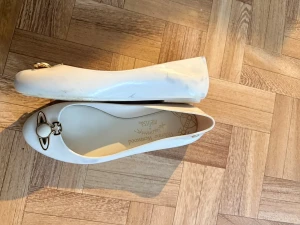 Vita ballerinaskor Vivienne Westwood - Säljer ett par vita ballerinaskor från Vivienne Westwood x Melissa med ikonisk guldfärgad orb-detalj framtill. Skorna är gjorda i mjuk plast och har rund tå samt låg klack. Perfekta för dig som gillar statementdetaljer och unik design. Dom har lite missfärgningar men det går nog bort, säljer pga de är för stora för mig❣️ det är storlek 38