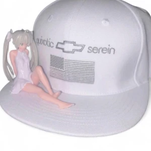 Vit keps Quixotic Serein brodyr - Säljer en vit snapback-keps från Quixotic Serein med broderad logga och text framtill samt en stilren flaggdetalj. Kepsen har platt skärm och justerbar baksida. Den har en figur från serien en anime serie.