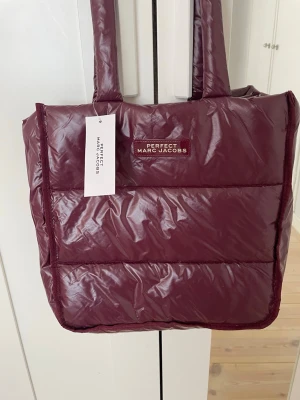Vinröd väska Marc Jacobs - Snygg vinröd shoppingväska från Marc Jacobs med quiltad design och vadderade handtag. Väskan har en glansig finish och logotyp framtill. Perfekt storlek för att bära med sig allt man behöver. Den går inte att köpa nånstans