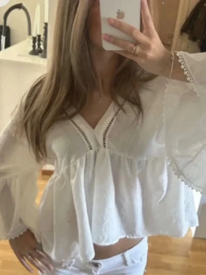 Vit bohemisk blus med v-ringning - Säljer en luftig vit blus i bohemisk stil med v-ringning och dekorativa spetsdetaljer längs halsen. Blusen har vida, långa ärmar och är tillverkad i ett tunt, lätt material som känns perfekt för sommaren. Passar till både jeans och kjol för en avslappnad look.