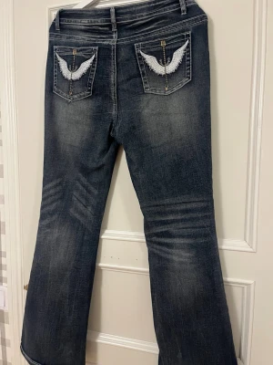 Mörkblå bootcut jeans med brodyr - Säljer ett par mörkblå bootcut jeans med slitningar och vita broderade vingar på bakfickorna. Jeansen har klassisk femficksmodell och kontrastsömmar. Perfekta för dig som gillar detaljer och en lite retro vibe.