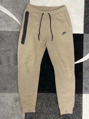 Beige Nike mjukisbyxor med svart detalj - Snygga beige mjukisbyxor från Nike med svart logga på höften och svart dragkedja längs ena fickan. Byxorna har ribbade muddar vid benslut, snörning i midjan och är tillverkade i mjukt material som ger en skön passform. Perfekta för chill eller träning.