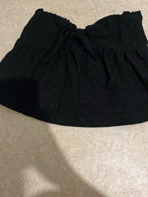 Svart bandeau topp med knut - Trendig svart bandeau topp med knut framtill och broderade håldetaljer. Toppen är axelbandslös och har en luftig passform, perfekt för sommardagar. Materialet känns lätt och bekvämt mot huden.